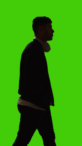 Vertikale-Videosilhouette-Eines-Mannes-Mit-Kabellosen-Kopfhörern,-Der-Vor-Einem-Grünen-Bildschirm-über-Den-Rahmen-Läuft